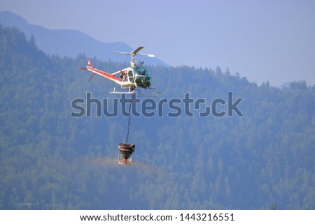 Profile view of a two man helicopter flying left to right while carrying fire retardant contained in a dumping bucket. 