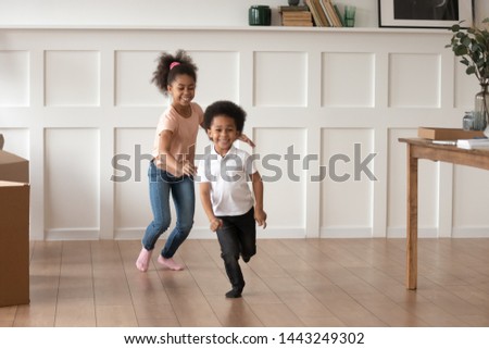 Excited smiling preschooler kids run in new empty home feel happy to move, overjoyed small brother and sister laugh have fun chasing each other playing in living room together. Entertainment concept Royalty-Free Stock Photo #1443249302