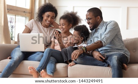 Happy african American young parents sit relax on couch with small kids watch funny video together, smiling black family with children rest on sofa with gadgets laugh enjoy cartoon on cellphone