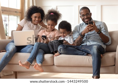 Happy young african American family with preschooler kids sit on couch use gadgets together, smiling black modern parents spend time with small children play on electronic devices. Technology concept