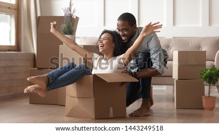 Overjoyed african American couple have fun feel euphoric moving together to new apartment, happy black husband ride smiling playful wife in cardboard box excited for relocation to own house Royalty-Free Stock Photo #1443249518