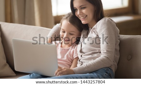Happy young mother and little daughter using laptop together, hugging, sitting on couch at home, smiling mum and adorable preschool girl looking at laptop screen, watching video or cartoons