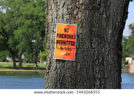 A shot of a bright and colorful orange sign that say's No Fireworks Permitted in this area. On tree's at Sterling Lake in Sterling Kansas USA. 
