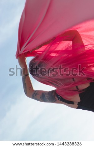 Pink-haired young woman with pieces of pink cloth dance near the field with wind generators . Windmill on sunset. Fashion portrait