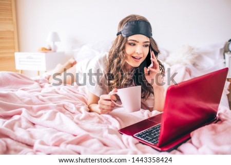 Young woman with dark hair. Talking on phone and looking on laptop screen. Cup in hand. Alone in bedroom. Lying on bed.