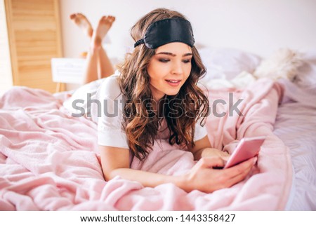 Young woman with dark hair lying on bed in bedroom alone. Holding phone in hands and looking at it. Device user.