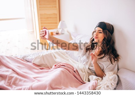 Young woman with dark hair taking selfie nd posing on phone camera. Alone in bedroom. Beautiful model in pajama. Morning daylight.