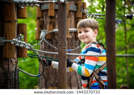 Young cute child boy in summer clothing, safety harness and helmet attached with carbine to cable moves slowly along rope way on green trees sunny bokeh background. Sport, game, leisure concept.
