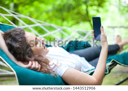 Summer day, Young woman lying in the hammock, with her hands behind his head, the other hand holding smart phone with blank screen. Woman uses a smartphone while lying in a hammock.