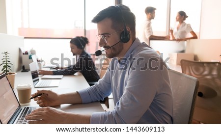Male professional call center telesales agent wear wireless headset using computer in customer care support service office with team, businessman operator telemarketer working on laptop at workplace Royalty-Free Stock Photo #1443609113