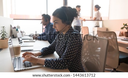 Serious young indian woman call center telesales agent operator telemarketer wear wireless headset work with team using computer in customer care service support helpline helpdesk multiethnic office Royalty-Free Stock Photo #1443613712