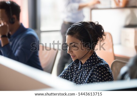 Focused indian female professional helpline call center agent operator telemarketer wear wireless headset work in customer care support service team office talking consulting client at workplace Royalty-Free Stock Photo #1443613715