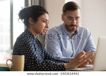 Focused male intern listening to serious indian business woman mentor teacher explaining online strategy looking at laptop computer teach trainee training new worker learning new skill at workplace Royalty-Free Stock Photo #1443616961
