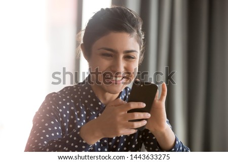 Happy indian ethnic business woman using smartphone at home office, smiling hindu girl having fun with cell phone texting sms play mobile games using apps for business or entertainment concept