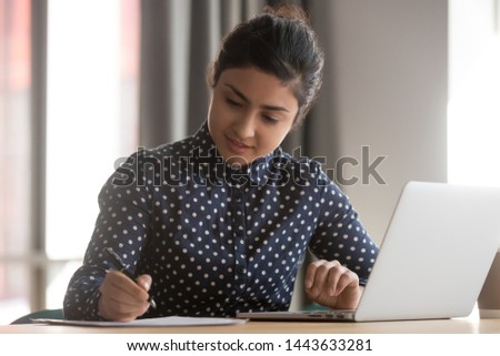 Young indian business woman student working studying online with laptop making notes sit at office desk, focused hindu female professional preparing report doing paperwork writing essay at workplace Royalty-Free Stock Photo #1443633281