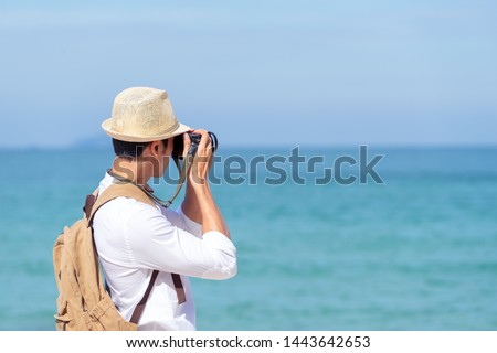 Candid of young attractive asian man hold camera taking photo in blue sky and sea view background. Happy asian hipster male photographer in youth freedom culture lifestyle travel in rear view concept.