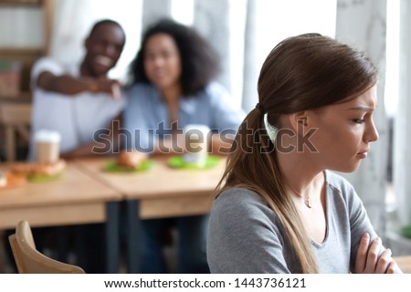 Upset frustrated young woman sitting apart from diverse people, suffering from bullying, mockery, gossip, offense, unfair attitude. Depressed female student excluded from bad friends company. Royalty-Free Stock Photo #1443736121