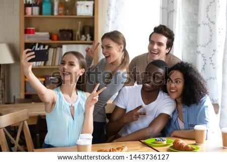 Happy young woman holding smartphone, making selfie or recording video with group of mixed race smiling female and male best friends, diverse people posing for photo with victory gesture in cafe.