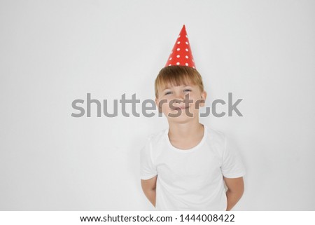 Isolated image of happy cute preteen boy with fair hair feeling shy holding hands behind back looking at camera and smiling, celebrating his birthday, wearing white t-shirt and red cone hat