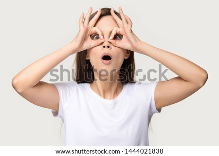 Nerdy young woman having fun making glasses shape with hands. Headshot of dummy millennial female do funny face with eyes wide open in studio isolated on grey background. People emotions concept