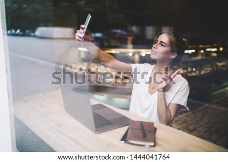 Positive female student making selfie photos via front camera on smartphone device in break of e learning online, hipster girl taking pictures for posting in social networks for attracting followers