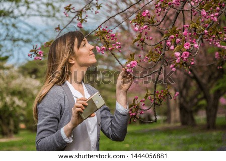 Woman holding a phone, smelling pink cherry blossom on a tree.