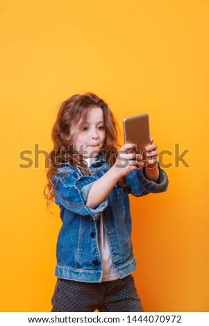 Concept of using modern technology by children. Close-up portrait of cute curly beautiful girl taking a self portrait on yellow background