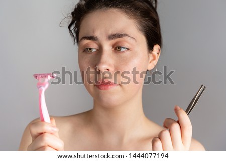 A young pretty woman holding a pair of tweezers and a razor, not knowing how to remove excess hair on her face.The concept of getting rid of unwanted hair