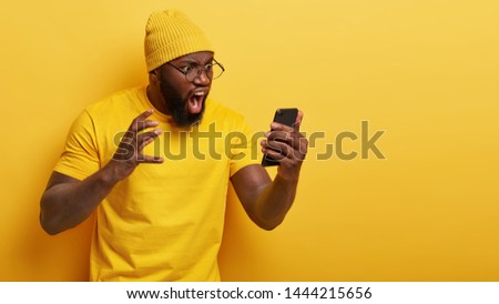 Outraged annoyed dark skinned man stares angrily at smart phone screen, shouts with irritation, dissatisfied with received message, has thick beard, wears casual yellow t shirt and headgear. Royalty-Free Stock Photo #1444215656