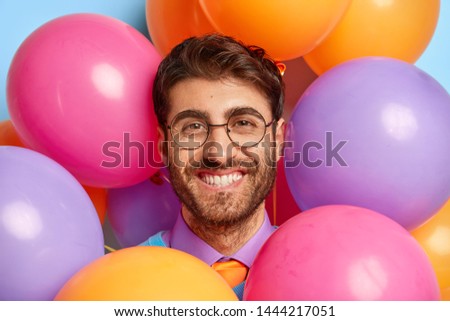 Birthday and celebration concept. Close up portrait of smiling man with bristle, wears spectacles for good vision, looks through colorful balloons, has party mood, makes photos before guests come