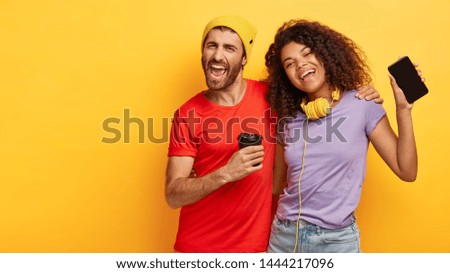 Horizontal view of playful positive mixed race couple cuddle and sing happily, boost energy and mood with cool music on smartphone, drink fresh coffee, look upbeat and entertained, isolated on yellow