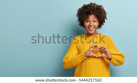 Pleasant looking smiling woman with Afro hairstyle makes heart gesture, confesses boyfriend in hearwarming feelings, shows love sign, wears oversized yellow jumper. Spread only love and care