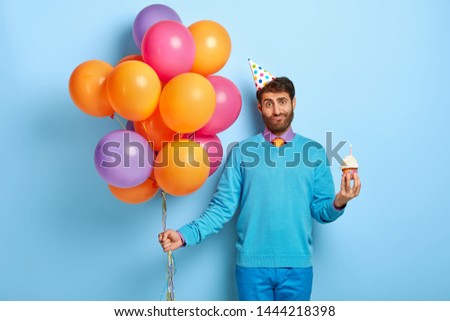 Horizontal shot of friendly guy with puzzled look, stands with cupckae and balloons, celebrates anniversary, has special occasion, wears party hat, blue jumper, anticipates for starting holiday