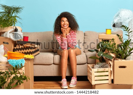 Delighted female apartment owner has dreamy expression, dressed in casual clothes, sits on sofa at cozy living room, carton boxes around, thinks about good future in own rental house, looks up