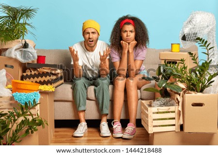 Horizontal shot of annoyed diverse married couple being busy with unpacking belongings, look in dissatisfaction, take break on sofa, pose in unfurnished apartment have to do repair being house tenants Royalty-Free Stock Photo #1444218488