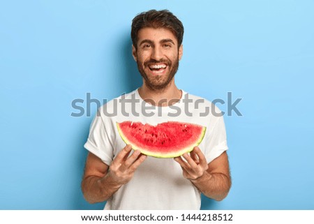 Waist up shot of smiling man enjoys summer day, holds slice of fresh ripe watermelon, wants to eat delicious fruit, wears casual white t shirt, isolated on blue background, has picnic during weekend