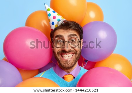 Headshot of handsome joyful birthday man celebrates anniversary in family circle, has perfect festive mood, receives congratulations and presents, wears cone paper hat, makes photo with balloons Royalty-Free Stock Photo #1444218521