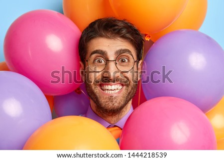 Headshot of handsome European man has broad smile, white perfect teeth, keeps face through helium multicolored balloons, has fun on birthday party, feels excited and glad. Celebration concept