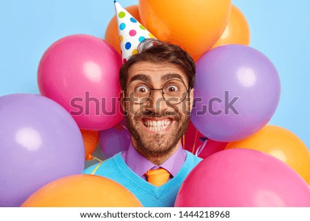 Positive student wears festive paper cap and spectacles, looks with brilliant smile, has leaving party after successfully passed final exams, stands through multicolored balloons, being in high spirit