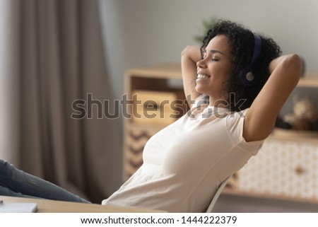 Happy african american girl wearing headphones relaxing listening to music taking break at work, calm smiling young black woman enjoying good lounge sound for stress relief sit at home office desk Royalty-Free Stock Photo #1444222379