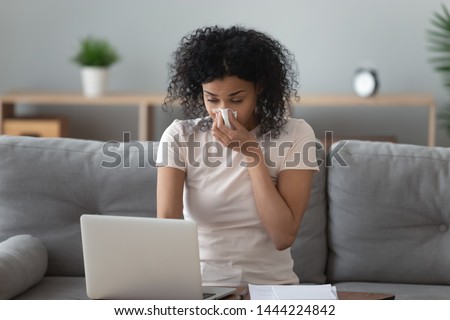 Allergic african woman blowing nose in tissue sit on sofa at home office study work on laptop, ill sick black girl got flu caught cold sneeze in tissue having allergy symptoms coughing holding napkin Royalty-Free Stock Photo #1444224842