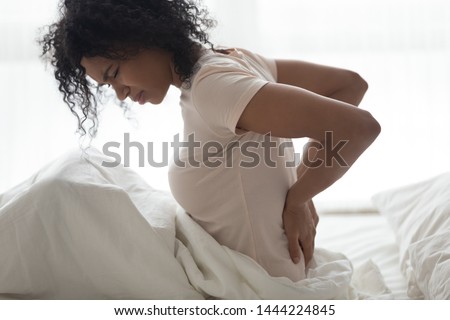 Sad young african woman touching back feeling backache morning discomfort low lumbar muscular kidney pain sit on bed after bad sleep waking up on uncomfortable mattress bending from backpain concept Royalty-Free Stock Photo #1444224845