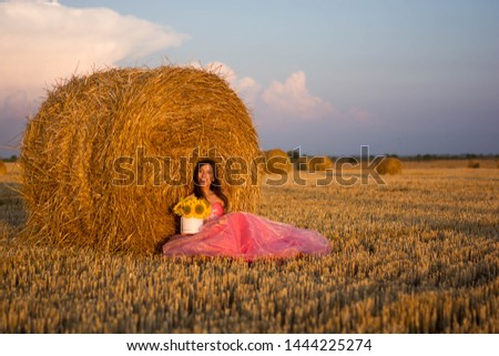 young beautiful woman smiling in a pink dress with flowers in her hands sitting near a hay style nature