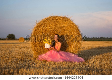 young beautiful woman smiling in a pink dress with flowers in her hands sitting near a hay style nature summer field