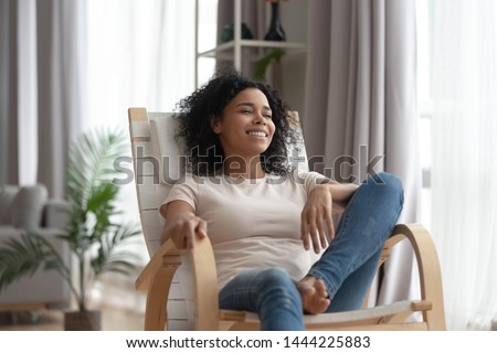 Smiling calm young black woman relaxing on comfortable wooden rocking chair in living room, happy healthy black girl enjoy breathing fresh air resting in armchair at home feel stress free on weekend Royalty-Free Stock Photo #1444225883