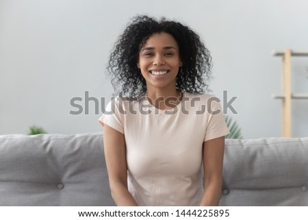 Smiling young african american woman sit on sofa looking at camera make video call or recording vlog at home, happy cheerful mixed race girl vlogger doing online chat shooting blog at home, portrait