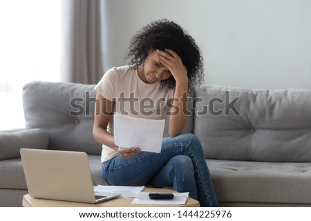 Stressed african woman holding papers calculate domestic bills at home worried about bankruptcy financial problem bank debt, depressed sad black lady frustrated about lack of finances sit on sofa