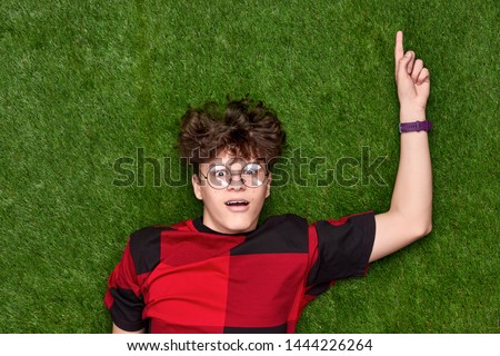 From above shocked youngster in nerdy glasses pointing up and looking at camera while lying on green lawn