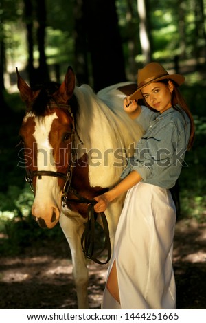 Young woman with horse in evening sunset light. Outdoor photography with fashion model girl. The girl and a horse are in the forest. Sunset. Sunrise. The model girl looks loke a cowboy. Wild West. The