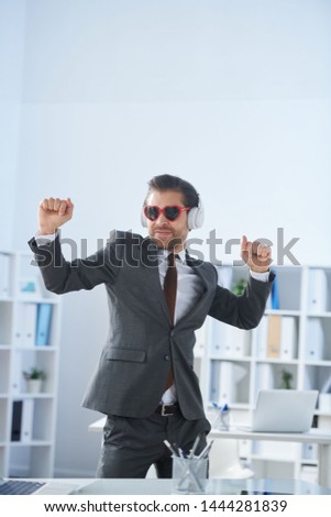 Happy man in formalwear, sunglasses and headphones dancing by workplace